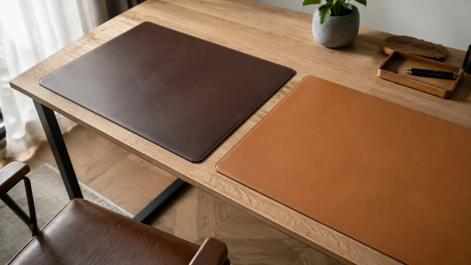 Two desk pads side by side on a wooden desk, one dark brown and one lighter tan, showing different edge stitching and surface textures