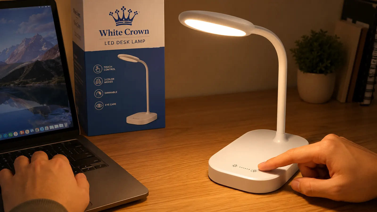 A hand reaching to press the touch dimmer on the White Crown LED desk lamp sitting on a wooden desk next to a laptop